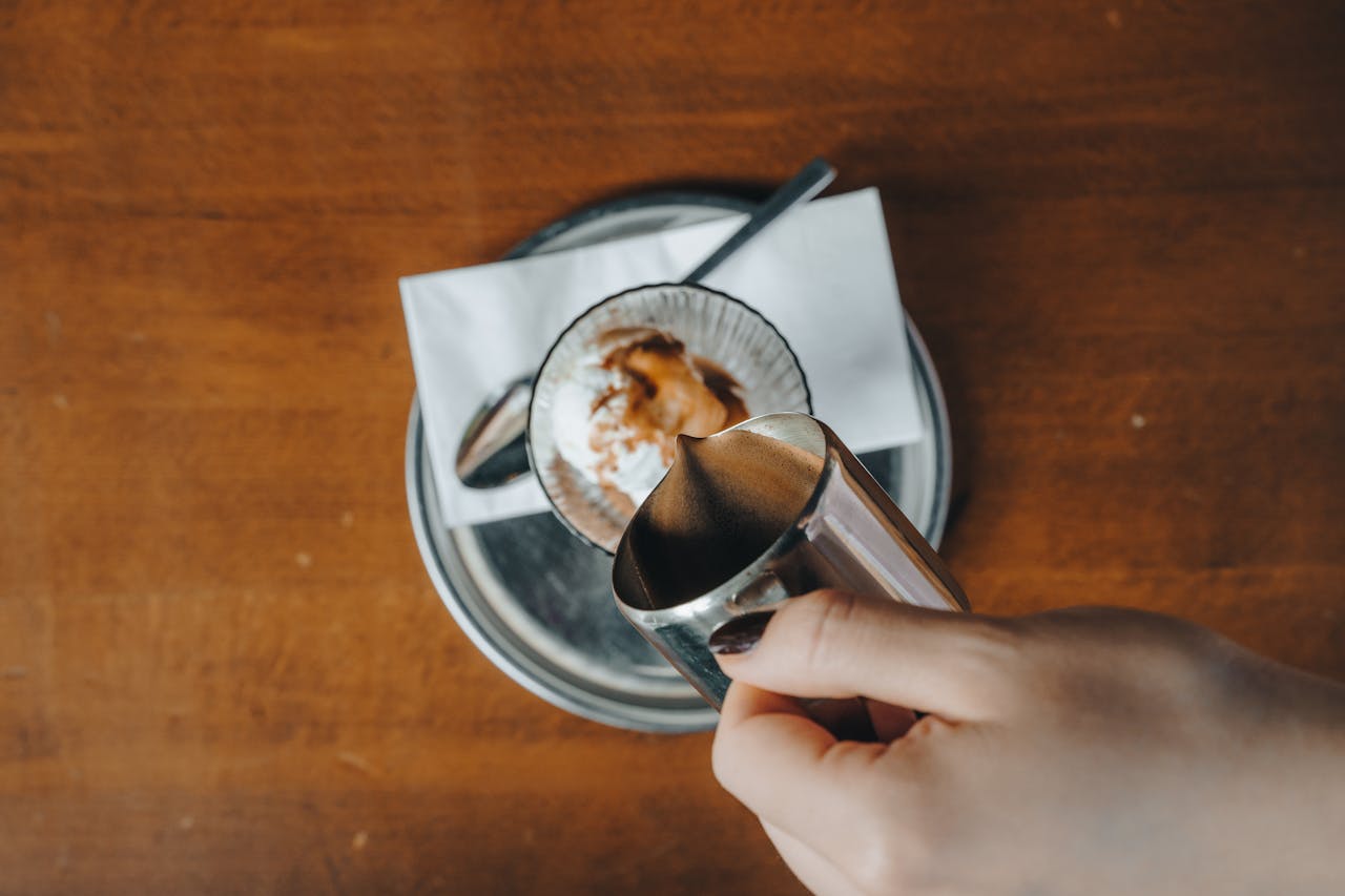 A high-angle view of hands pouring coffee into a cup with a spoon and napkin on a wooden table.