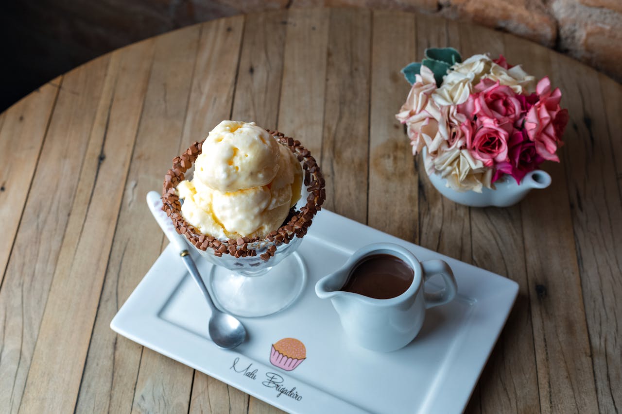 Scoops of ice cream served with chocolate sauce on a wooden table, alongside decorative flowers.